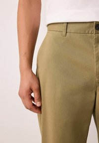 Khaki nohavice s presným strihom a hladkou textúrou, s gombíkovým zapínaním a bočnými vreckami. Tkanina je jemne textúrovaná.