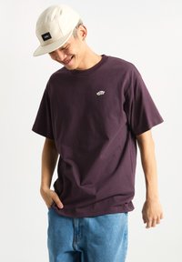 Camiseta de algodón morada con mangas cortas, cuello redondo y un pequeño logotipo gráfico en el pecho. Se lleva con pantalones de mezclilla azul claro.