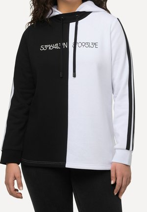 Person iført en sort og hvid opdelt hættetrøje med trykte koordinater på brystet og sorte striber langs ærmerne.