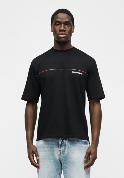 Νέος άνδρας που φοράει μαύρο μπλουζάκι Dsquared2 με λεπτή πορτοκαλί γραμμή στο στήθος και ανοιχτόχρωμο ξεβαμμένο τζιν, στέκεται μπροστά σε λευκό φόντο.