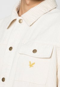 White corduroy jacket featuring a structured collar, brass snap buttons, and a small yellow bird embroidery on the front pocket.