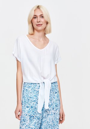 Biała bluzka z krótkim rękawem i dekoltem w kształcie litery V, z przodu z wiązaniem, w zestawieniu z niebieskimi, wzorzystymi spodniami o szerokich nogawkach z kwiatowymi motywami.