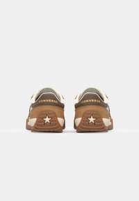 Pár hnědých a krémových tenisek Converse zobrazených zezadu, s důrazem na hvězdný emblém na patě a nápis „CONVERSE“ nad ním.