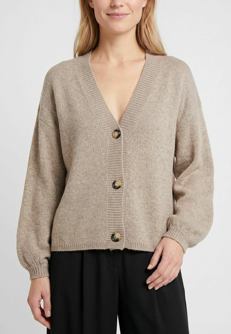 Femme portant un cardigan en maille beige avec trois boutons et manches longues, associé à un pantalon noir ample, sur un fond uni.