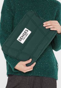 Gepolstertes, dunkelgrünes Laptop Sleeve mit einem weißen Logo-Patch, ausgestattet mit einem Reißverschlussverschluss und einem gepolsterten Design zum Schutz.