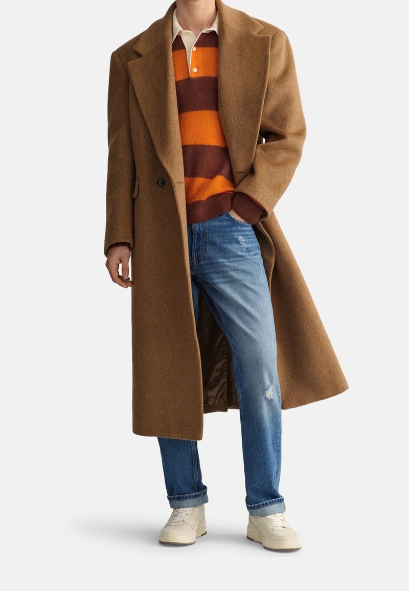 Manteau en laine marron associé à un pull rayé orange et marron, jean bleu et baskets crème, présentant un look décontracté en couches.