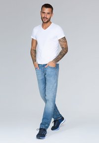 Camiseta blanca de cuello en V, jeans de mezclilla azul claros y zapatillas deportivas negras con acentos azules. Hombre con tatuajes en los brazos, de pie con las manos en los bolsillos.