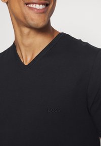 T-shirt noir à col en V en tissu lisse, avec un logo en relief subtil sur la poitrine, design ajusté avec des manches courtes.