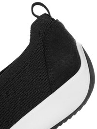 Primer plano de una zapatilla negra sin cordones con parte superior de punto, talón de ante, lengüeta para tirar y suela de goma blanca.