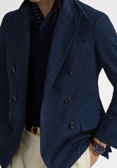 Tamsiai mėlynas dvieilis švarkas su aštriomis atlapais, tekstūruota medžiaga, dvi didelės sagos ir kišenė su lopu. Dėvimas ant tamsios megztos marškinėlių.