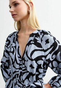 Helle blaue Bluse mit kräftigem schwarzen Blumenmuster, V-Ausschnitt, Knopfverschluss und gerafften Ärmeln. Weicher, strukturierter Stoff.