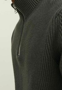 Primo piano di una persona che indossa un pullover a costine grigio scuro con una zip argentata e un colletto alto parzialmente zipato.