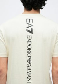 Sötét göndör hajú személy krémszínű pólóban, melynek hátulján függőlegesen fekete "EA Emporio Armani" logó látható.