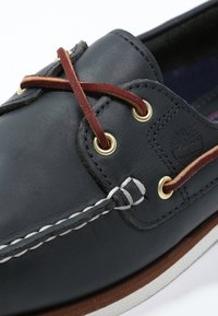 Chaussure bateau en cuir bleu marine avec des lacets marron et des œillets dorés. Caractérisée par une semelle en caoutchouc blanche et des coutures contrastantes le long du bord.