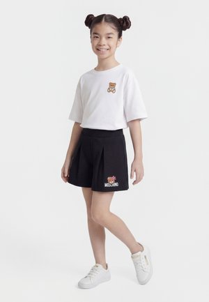 Młoda dziewczyna z dwoma kokami, ubrana w białą koszulkę z misiem, czarną plisowaną spódnicę Moschino oraz białe sneakersy Moschino, uśmiechająca się.