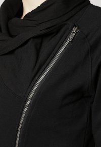 Svart zip-jacka med en snug krage och mjukt tyg, med en framträdande silvrig dragkedjedetalj på sidan.