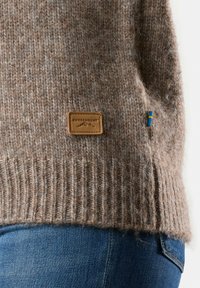 Maglione marrone lavorato a maglia con una superficie testurizzata, orlo a costine e un'etichetta in pelle con la scritta "SWEDEMOUNT." Piccola bandiera con accenti blu e gialli.