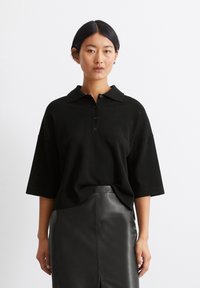 Polo en coton noir à manches courtes, avec une patte de boutonnage et une coupe décontractée, associé à une jupe en cuir noir. Texture lisse, design minimal.
