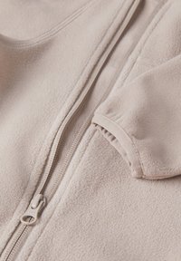Helle beige Fleecejacke mit einem Reißverschluss an der Vorderseite, gerippten Bündchen und weicher Textur. Sichtbare Nähte entlang der Kanten und des Reißverschlusses.