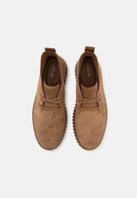 Scarpe casual in suede marrone con lacci e suole in gomma, viste dall'alto su uno sfondo bianco, marchiate "Clarks" all'interno.