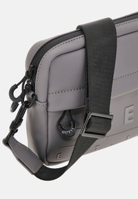 Bolso cruzado sintético gris con acabado texturizado, correa negra y cierre de cremallera. Presenta un detalle de logo y acentos de hardware negro.