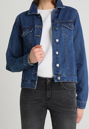 Osoba ubrana w niebieską kurtkę dżinsową na białej teksturowanej koszuli i czarnych dżinsach, z lewą ręką trzymającą klapę kurtki.