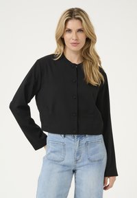 Schwarze cropped Jacke mit Rundhalsausschnitt, vorderem Knopfverschluss und langen Ärmeln, kombiniert mit hellblauen hoch taillierten Jeans.