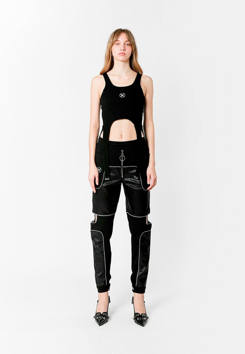 Top corto negro con recortes, que presenta un logo. Pantalones negros con bolsillos con cremallera, detalles en cuero y ribetes blancos. Tacones negros de punta.