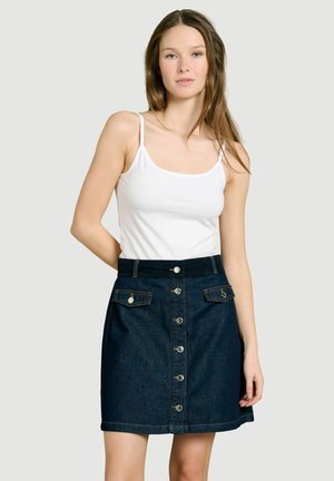 Jeune femme aux longs cheveux bruns portant un haut blanc sans manches et une jupe en denim foncé boutonnée, sur un fond clair uni.