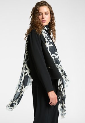 Donna con capelli ricci che indossa un completo nero e una sciarpa con motivo bianco e nero drappeggiata sulle spalle, su uno sfondo semplice.