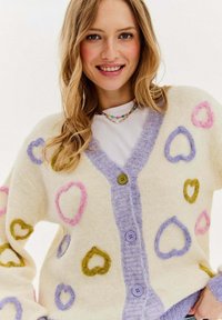 Pletený kardigan s motivy srdce v růžové, fialové a zelené barvě na krémovém pozadí. Má levandulové zapínání vpředu a strukturovaný povrch.