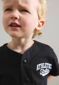 Svart kortärmad skjorta med rund halsringning, med vit text "ATHLETIC STATE" och knappdetaljer framtill. Mjuk tygkänsla.