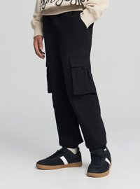 Czarne spodnie cargo z dużymi bocznymi kieszeniami, o prostym kroju, wykonane z teksturowanej tkaniny, zestawione z czarnymi trampkami z białymi akcentami.