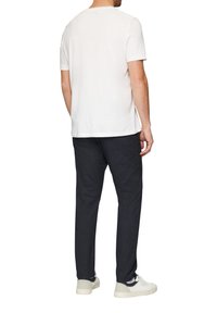 Witte t-shirt met korte mouwen, gecombineerd met donkere broeken, gedragen met lichte sneakers. De achtergrond is eenvoudig wit, wat de kleding benadrukt.