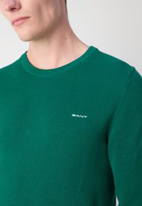 Pulover tricotat verde, realizat dintr-un material texturat, cu guler rotund și un mic logo alb GANT pe piept.