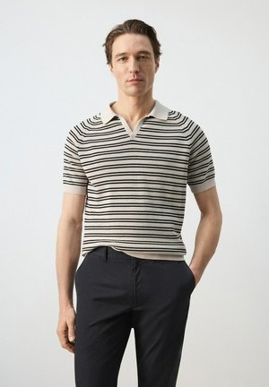 Uomo che indossa una polo beige a maniche corte con strisce orizzontali nere e pantaloni scuri, in piedi con una mano in tasca.