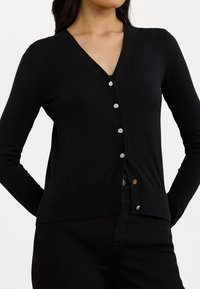 Svart v-ringad kavaj med långa ärmar, med silverknappar framtill och en texturerad yta. Bärs över mörka byxor.