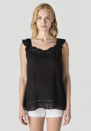 Mujer joven con cabello largo y rubio, vestida con una blusa sin mangas de encaje negro y pantalones cortos blancos, de pie frente a un fondo gris liso.