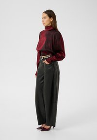 Blusa di raso bordeaux con colletto alto, abbinata a pantaloni in pelle nera a gamba larga e décolleté appuntiti color marsala. La cintura è decorata con borchie argentate.