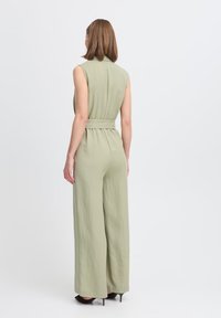 Salopette verde senza maniche con vita cinturata, gambe larghe e tessuto strutturato. La vista posteriore mostra una silhouette liscia e dettagli minimi.