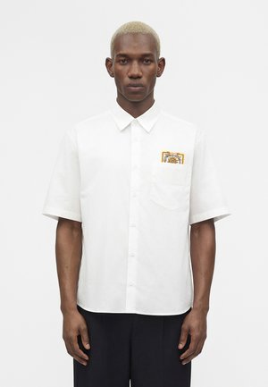 Hombre con cabello corto y rubio que lleva una camisa blanca de manga corta abotonada con un parche decorativo en el bolsillo del pecho.