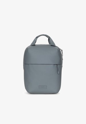 Mochila rectangular gris con asa superior, bolsillo frontal con cremallera y superficie lisa, de pie sobre un fondo blanco.