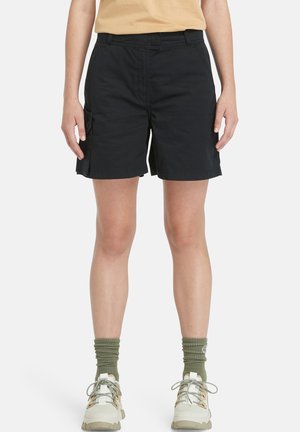 Pantaloni cargo neri in cotone con tasche frontali e laterali, caratterizzati da una vestibilità rilassata e un design lungo fino al ginocchio, abbinati a scarpe leggere e calzini verdi.