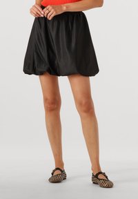 Zwarte satijnen pofrok met een elastische tailleband, met een gladde textuur en een ronde silhouet. Gecombineerd met geweven zwarte en tan schoenen.