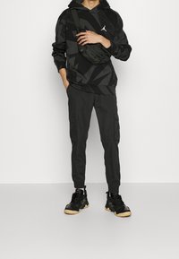 Hoodie noir à motifs avec une poche kangourou, associé à un pantalon cargo noir et des baskets noires avec des semelles en gomme. Un sac à bandoulière compact est porté.