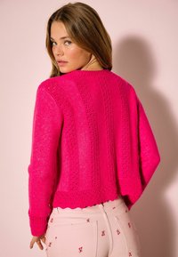 Pulover tricotat roz aprins cu corp texturat, prevăzut cu dungi verticale și tiv ondulat. Material ușor, asortat cu pantaloni deschiși la culoare cu model.