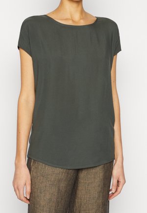 Frau trägt eine dunkelolivfarbene Bluse mit kurzen Ärmeln und braune, strukturierte, hoch taillierte Hosen, steht vor einem schlichten, hellen Hintergrund.