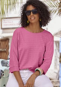 Rosa strukturierte Bluse mit rundem Halsausschnitt und Dreiviertelärmeln, kombiniert mit weißen Hosen. Auffälliges, mehrfarbiges Armreifen am Handgelenk.