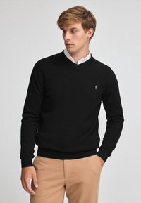 Jersey negro de punto con cuello en V, puños y dobladillo de canalé; presenta un pequeño logo bordado en el lado izquierdo del pecho. Lleva encima una camisa blanca con cuello.