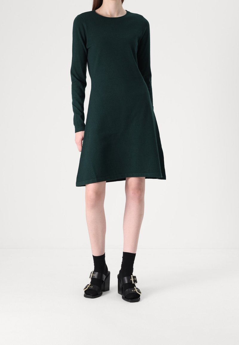 Femme portant une robe vert foncé à manches longues, longueur genou, avec des chaussettes noires et des sandales noires à talons avec boucles, debout sur un fond uni.
